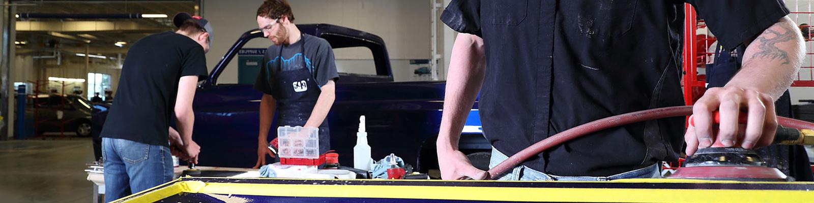 Student in shop shirt buffing tailgate in collision lab, others working in background