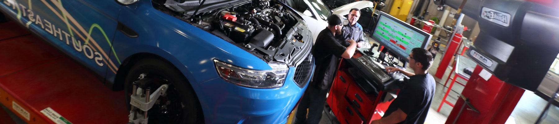 Three men in front of a computer screen next to a blue sedan on a lift with the hood open. 